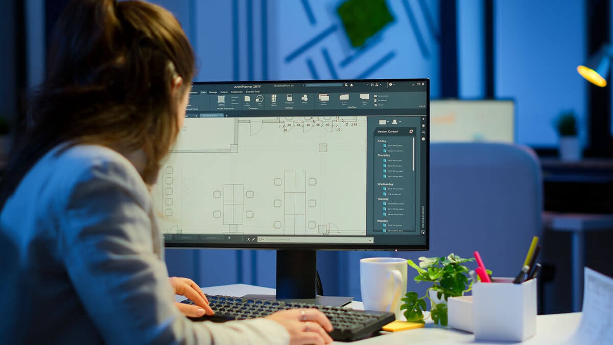 Ansicht von hinten: Eine Frau sitzt in einem Büro an einem Schreibtisch mit PC, die Hände auf Tastatur und Mouse und bearbeitet einen Grundriss, der auf dem Monitor zu sehen ist.