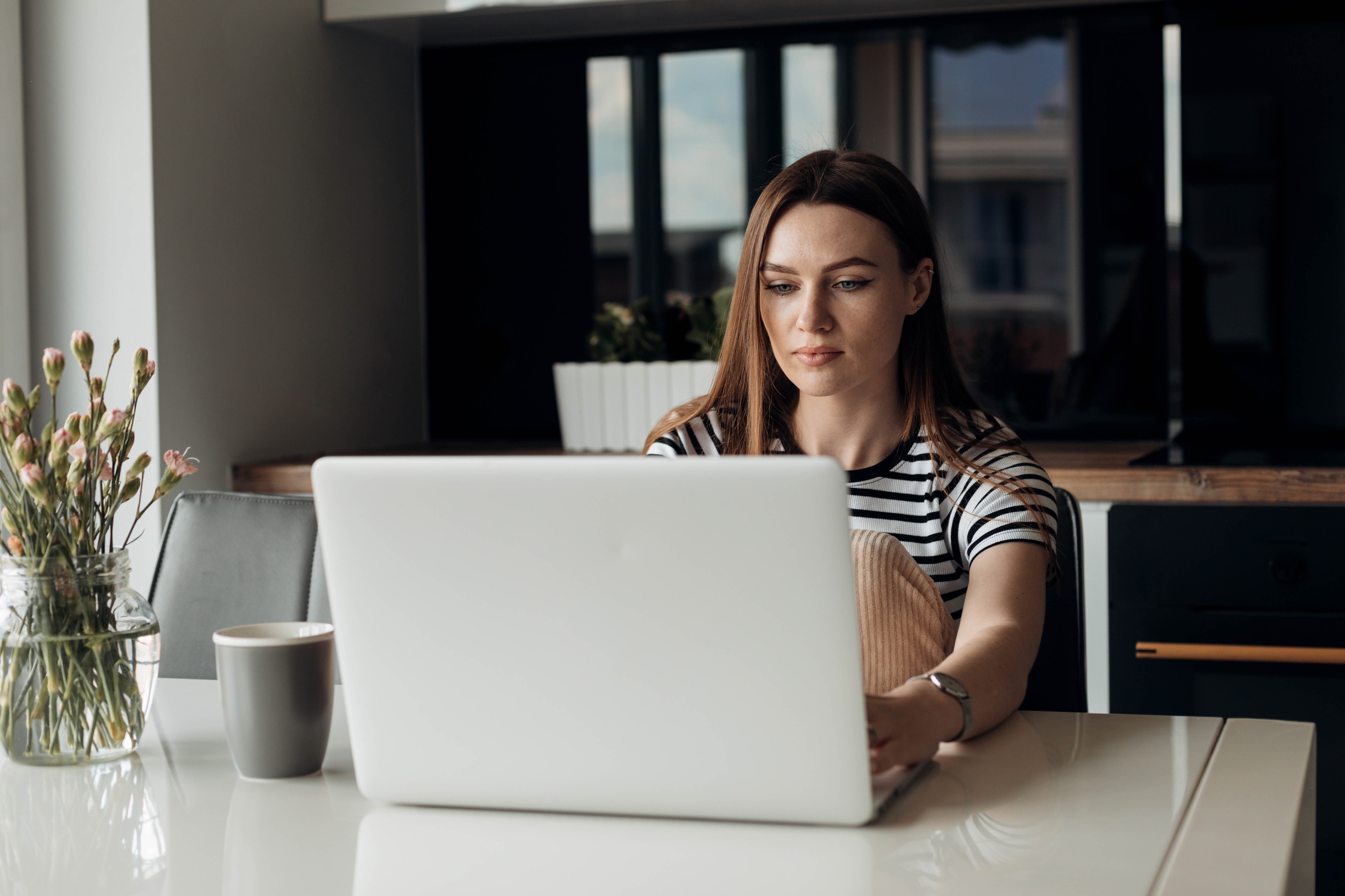 Eine Frau sitzt an einem Küchentisch und arbeitet an einem Laptop im Hintergrund sind dunkle Küchenschränke zu sehen.