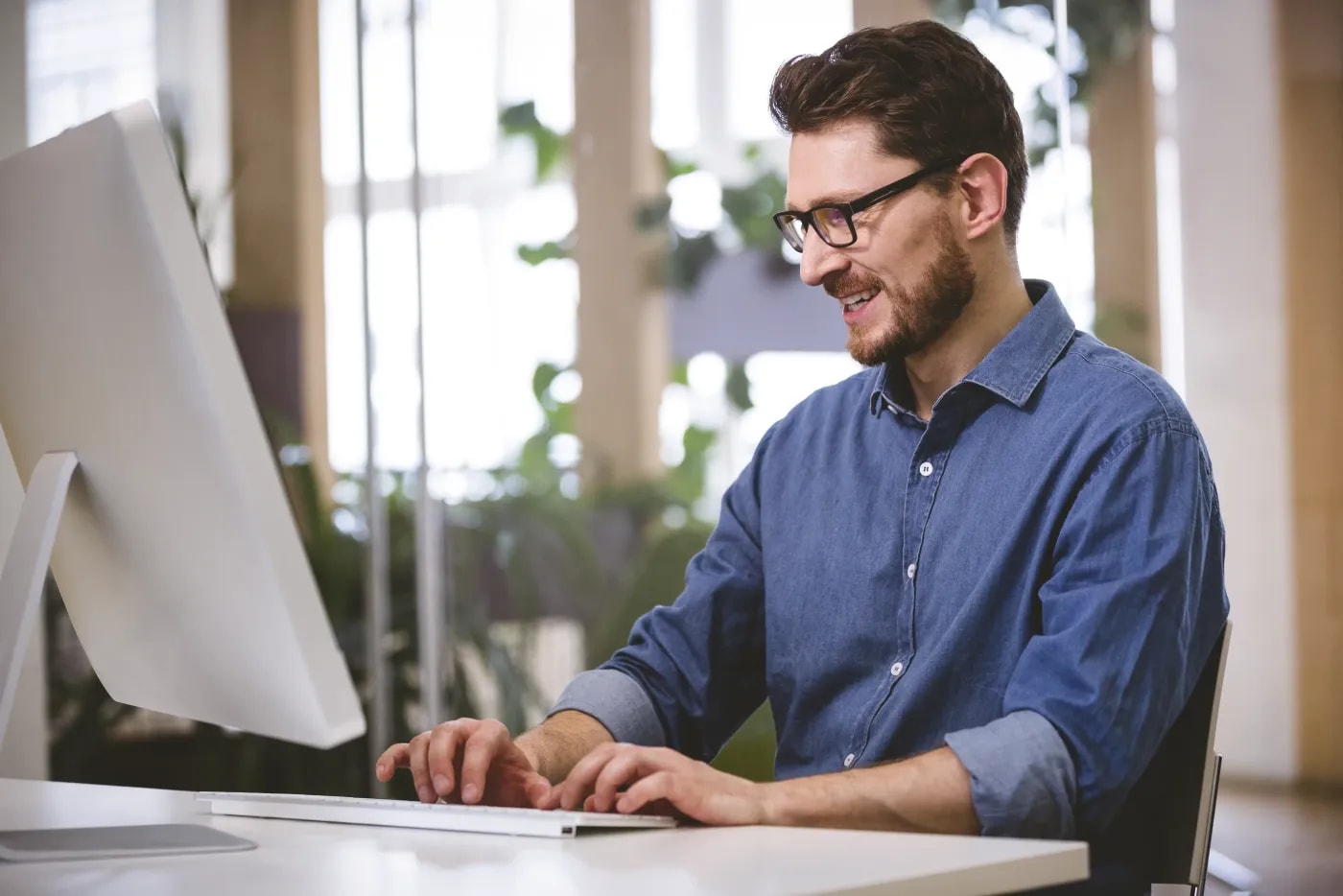 Ein Mann mit blauem Hemd und Brille sitzt in einem Büro mit Glastüren und arbeitet lächelnd an einem PC.