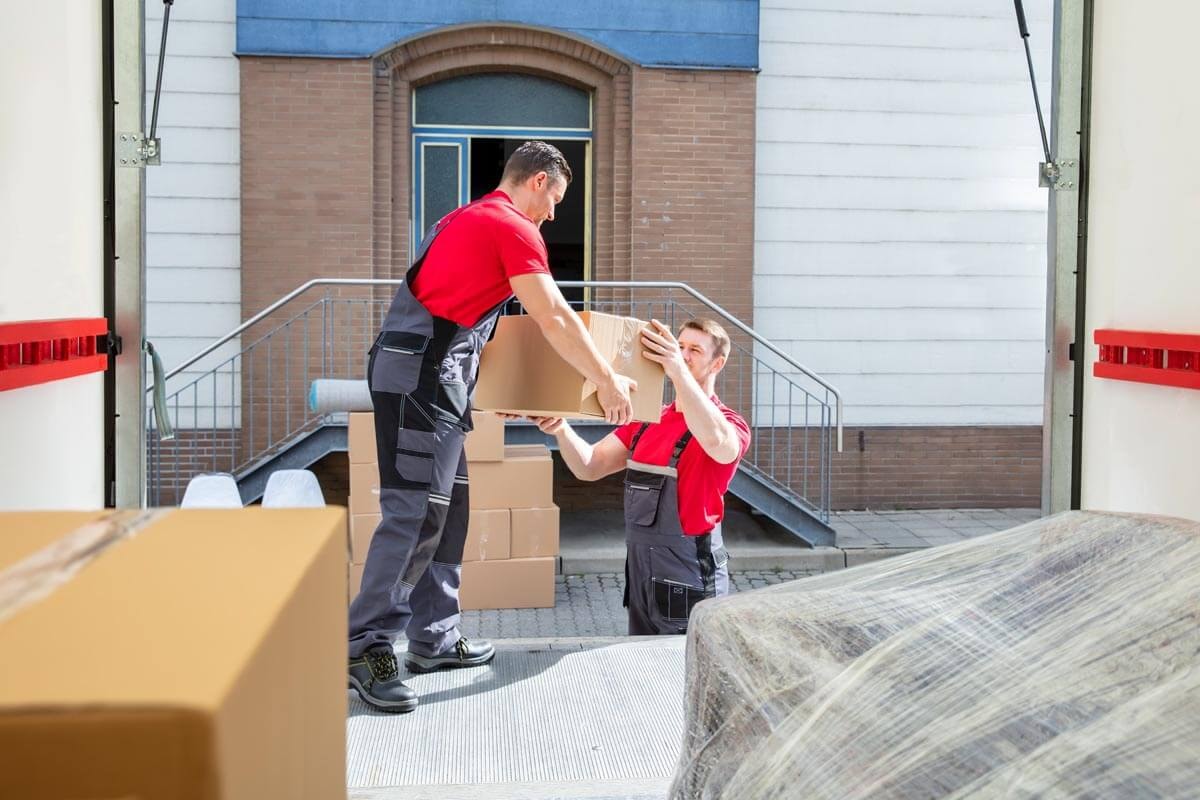 Zwei Umzugshelfer in roter Arbeitskleidung reichen sich aus dem geöffneten Laderaum eines Umzugstransporters Kartons heraus; im Hintergrund ein Backsteingebäude mit Treppenanlage.