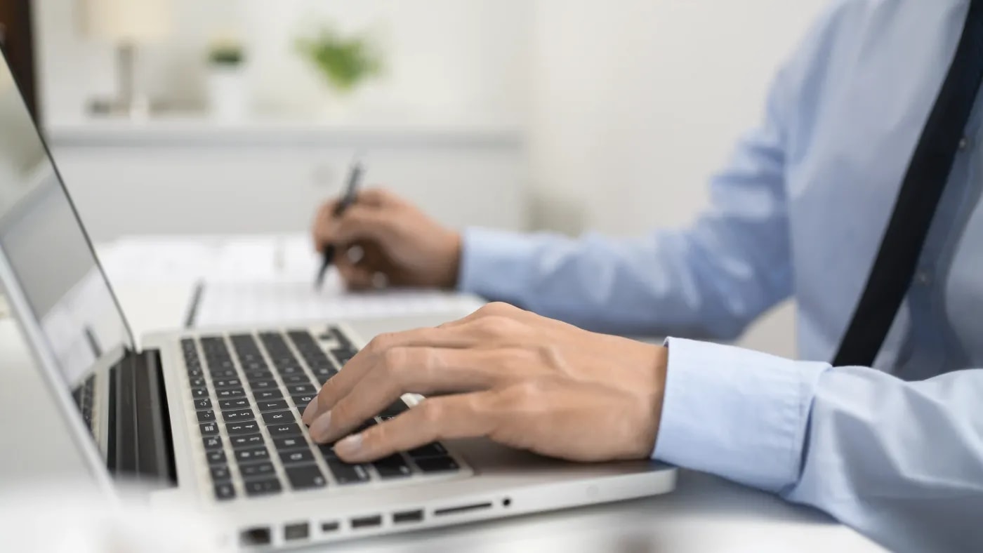 Eine Person sitzt am Schreibtisch vor einem Laptop und überträgt Daten von einem Blatt neben sich in den Laptop.