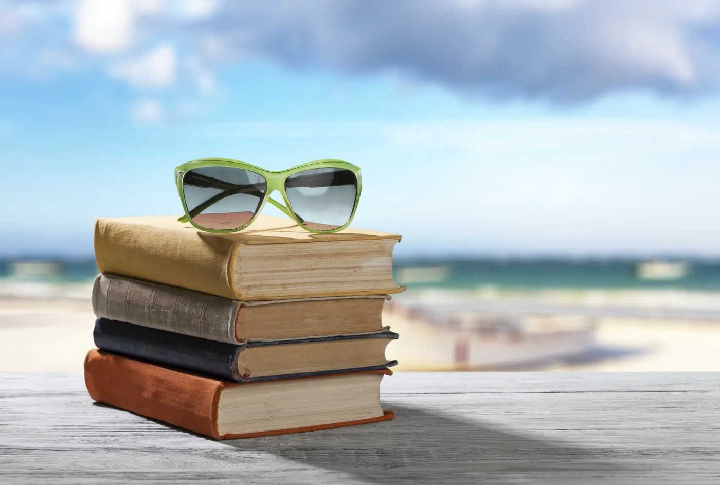 Ein Stapel mit vier Büchern mit Sonnenbrille oben drauf liegt auf einer Holzplatte vor einem verschwommenen Strandpanorama im Hintergrund.