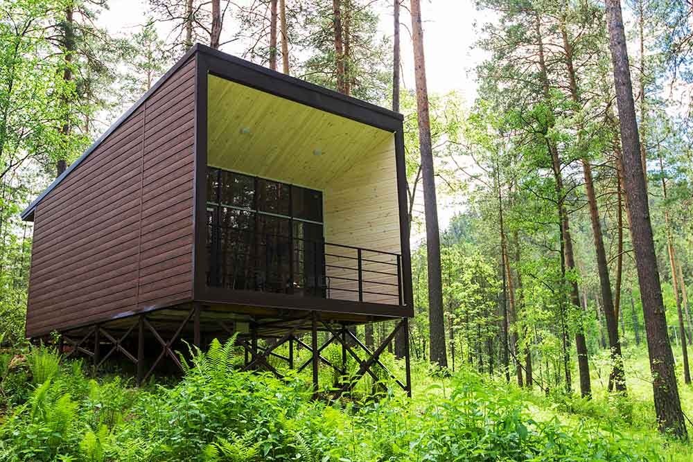 Kubistischer Bungalowaus Holzlatten auf einem Stahlgestell an einem dichtbewachsenen Steilhang im Wald.
