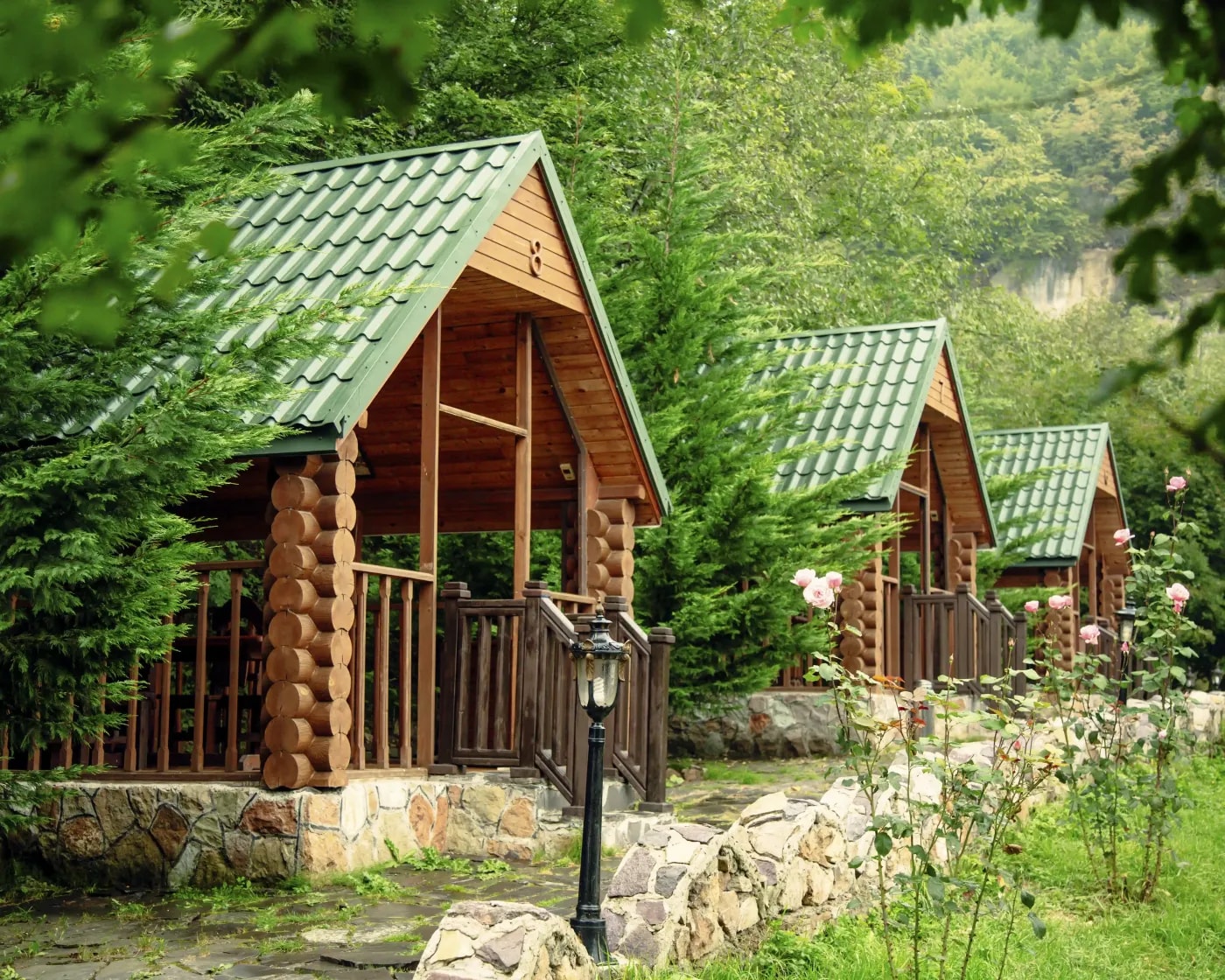 Frontansicht von drei baugleichen Bungalows mit grünem Satteldach zwischen Tannen mit Felsberghang im Hintergrund.