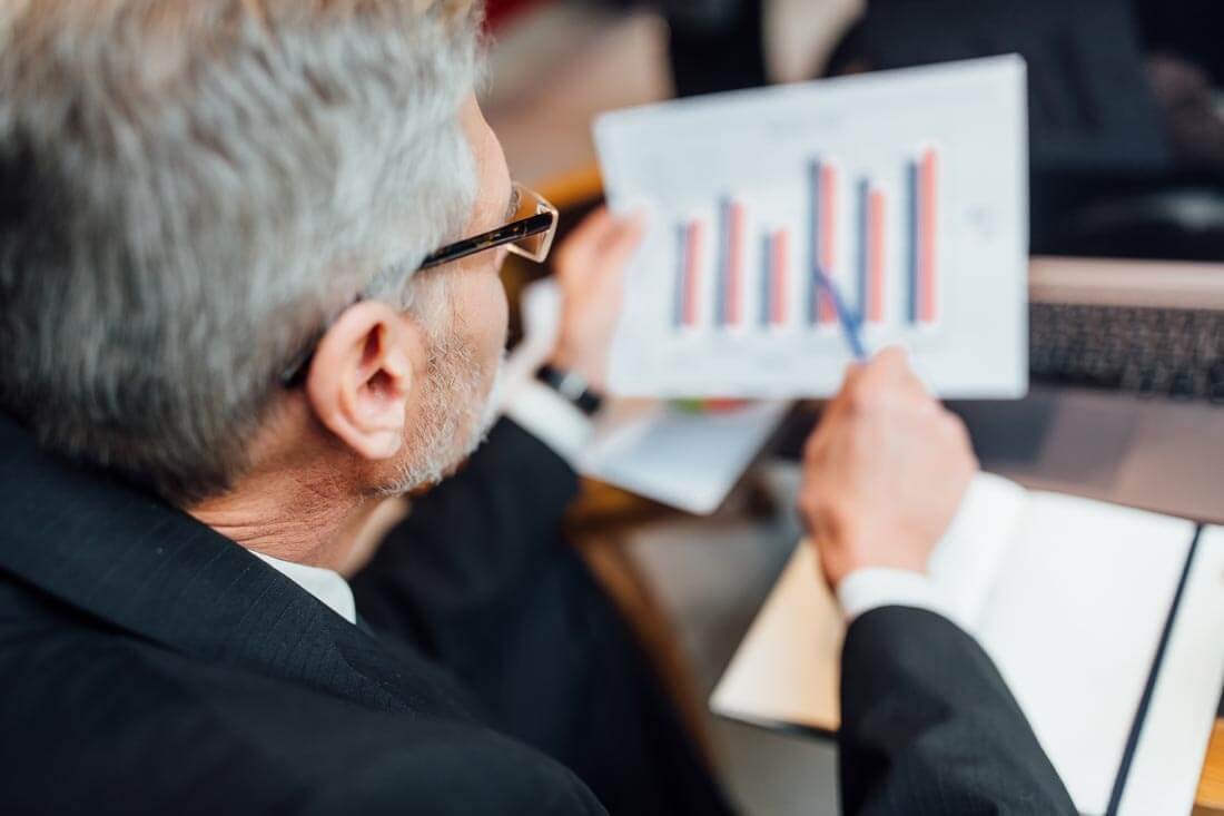 Blick über die Schulter eines älteren Investmenbankers, der an einem Konferenztisch sitzt und ein Balkendiagram durchgeht.