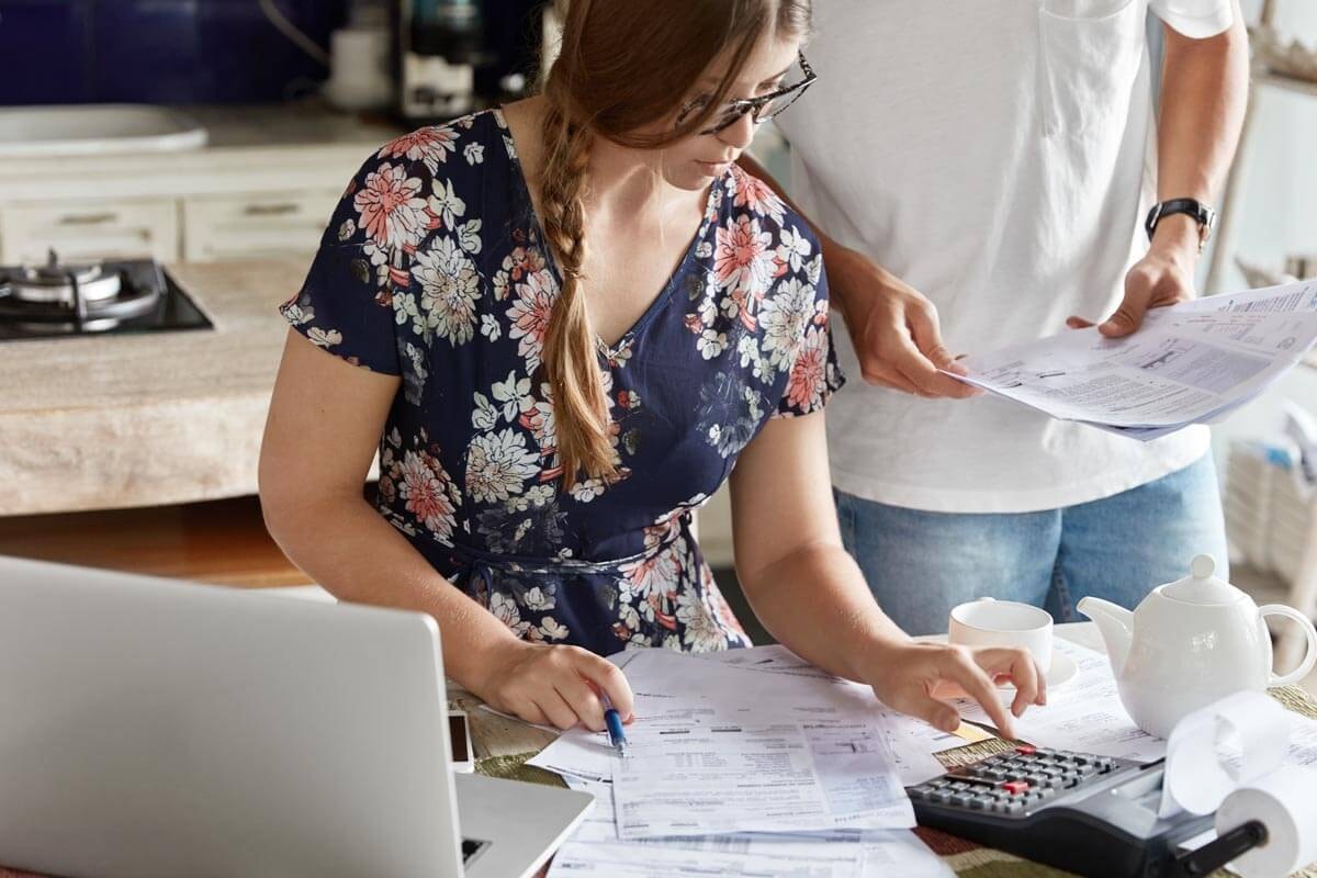Eine Frau sitzt an einem Schreibtisch und stellt Berechnungen mit Hilfe eines Taschenrechners an deren Informationen sie aus einem Stapel Unterlagen vor ihr auf dem Tisch entnimmt, während ein Mann neben ihr steht und weitere Unterlagen bereithält.