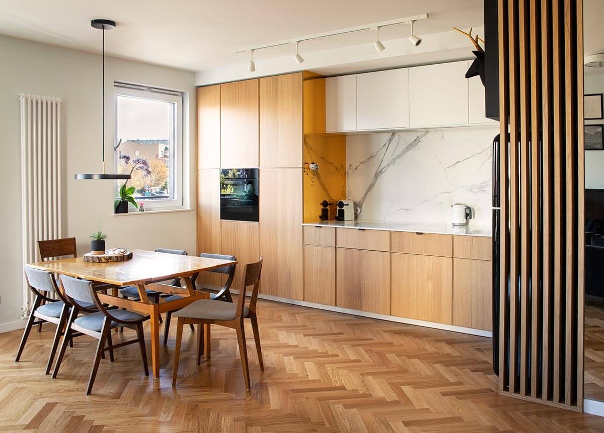 Blick von der Seite auf eine offene Küche mit Küchenzeile in Eichenoptik und heller Arbeitsplatte, mit Esstisch davor mittig im Raum.