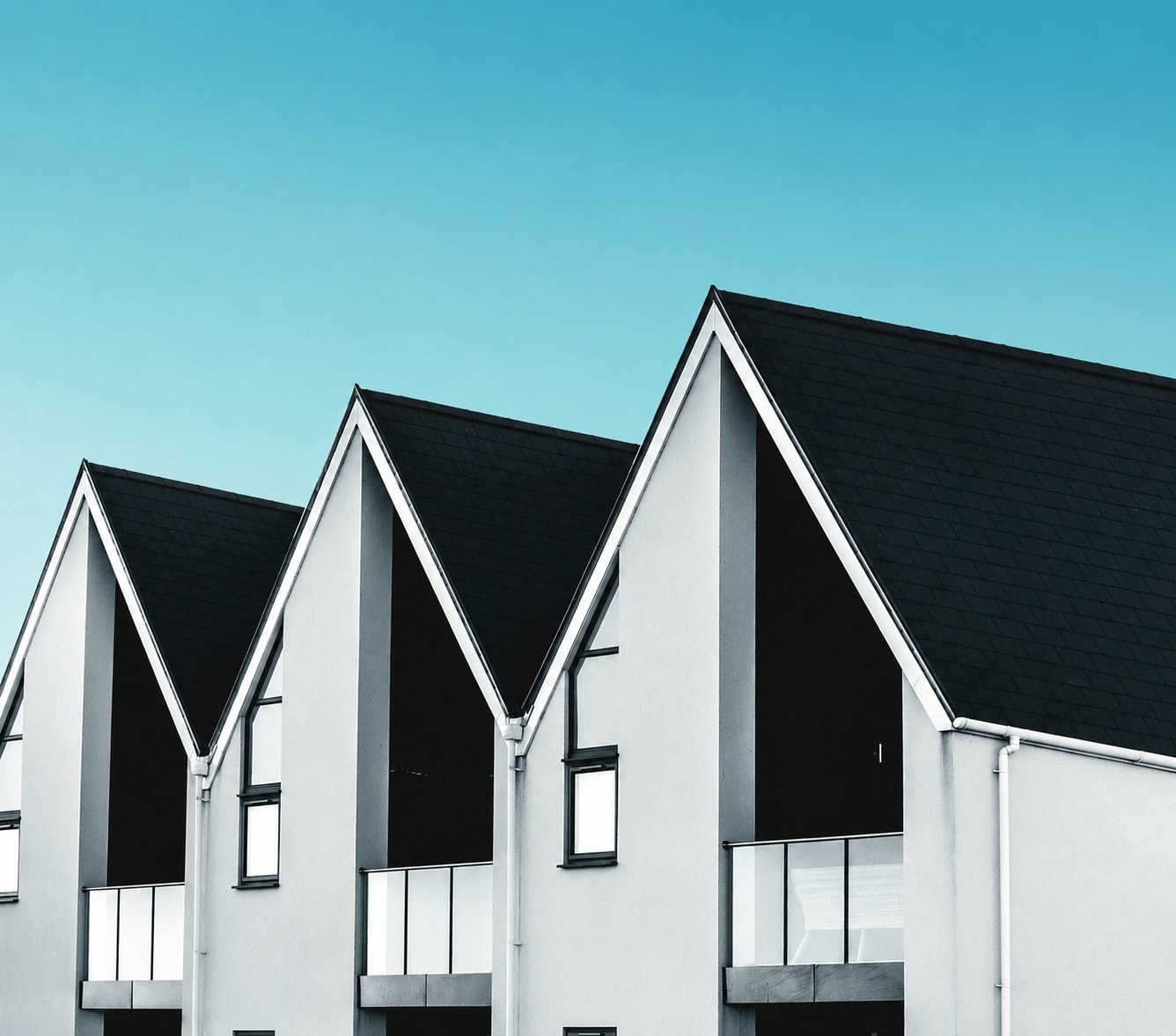 Modernes Reihenhaus mit zurückgesetzten Balkonen mit Glasplatten-Geländer in der geometrisch designten Fassade und steilem Satteldach mit schwarzen Schindeln vor einem blauen Himmel.