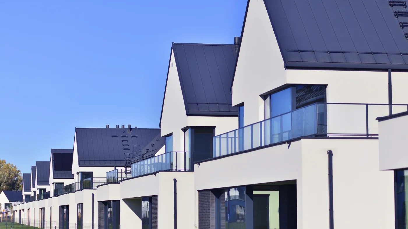 Modernes Reihenhaus mit hellem Putz und schwarzen Dachplatten, Balkonen mit verglastem Geländer und tiefblauem Himmel darüber.