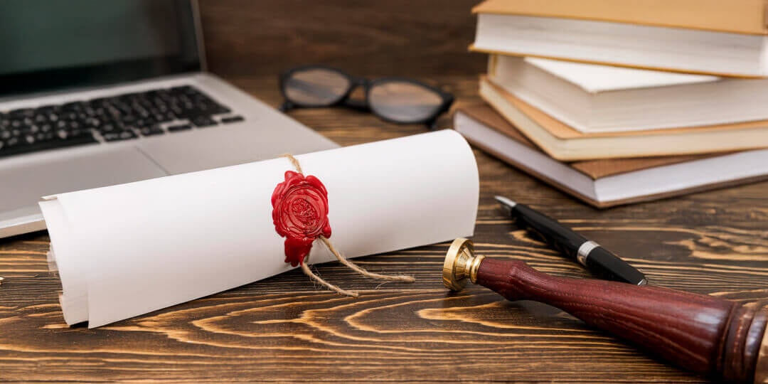 Gerollte Urkunde mit rotem Wachssiegel auf einem Holztisch, daneben Bücherstapel, Brille, Laptop und ein Siegelstempel.